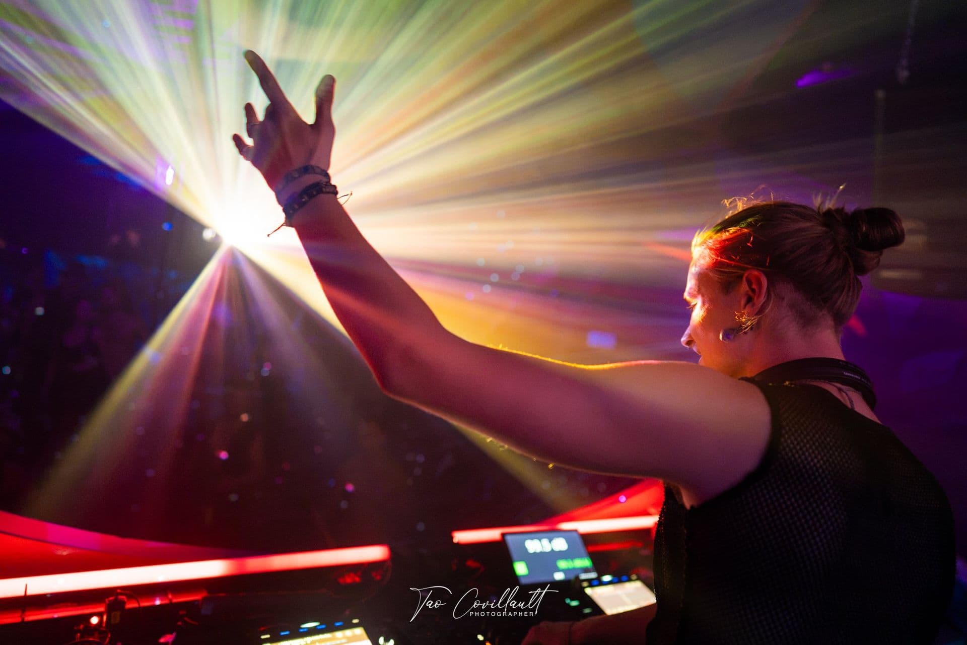 A DJ reaching up into a burst of stage light behind the booth