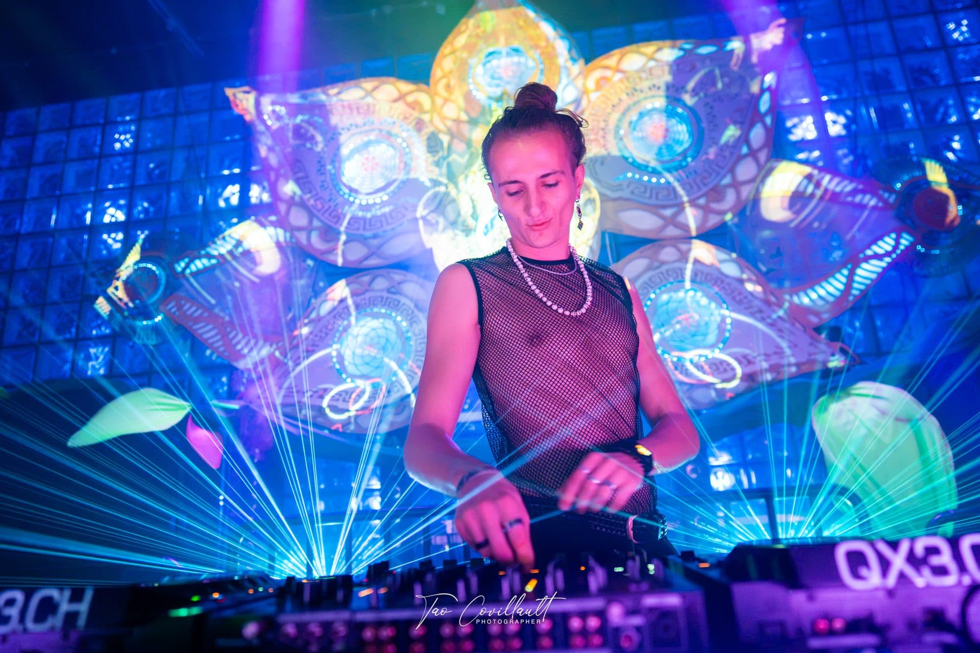 A DJ at the decks in front of a glowing lotus installation, framed by a fan of laser beams